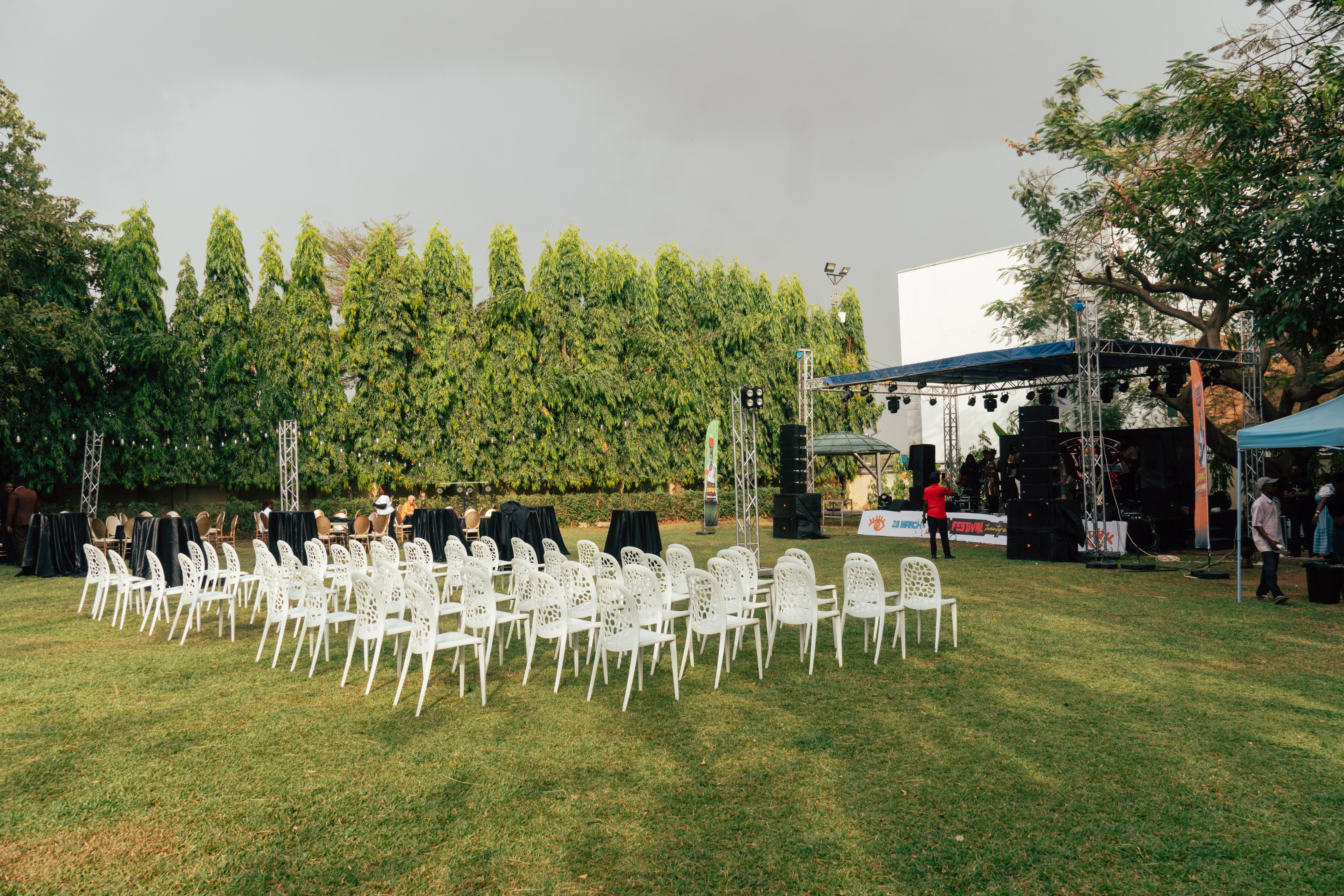 Outdoor event venue with white chairs, stage equipment, and surrounding greenery.