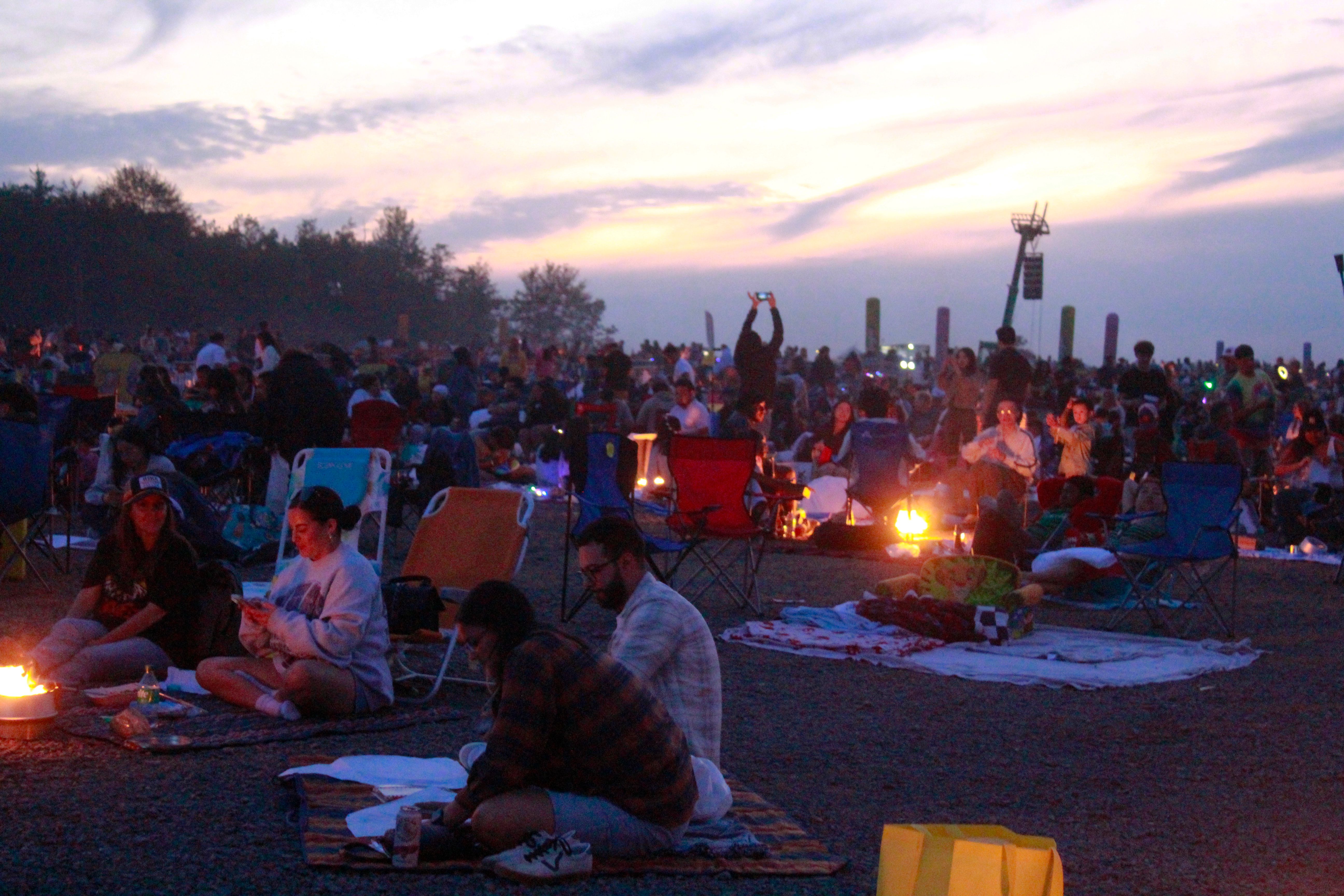 Music festival guests relaxing in camping chairs around fire pits at sunset.